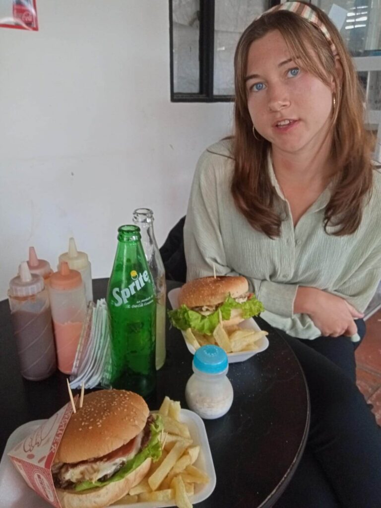 Ik zit aan tafel en kijk naar de camera. Op de tafel staan 2 borden met op elk bord een burger