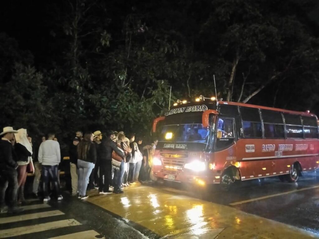 Het is avond en donker. Er staat een rode bus langs de weg. Er staan zo'n 15 mensen in de rij om in de bus in te stappen