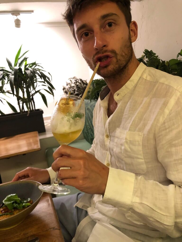 Niels heeft een drankje in z'n hand en het rietje van het drankje in z'n mond. Hij kijkt in de camera terwijl hij een slok neemt