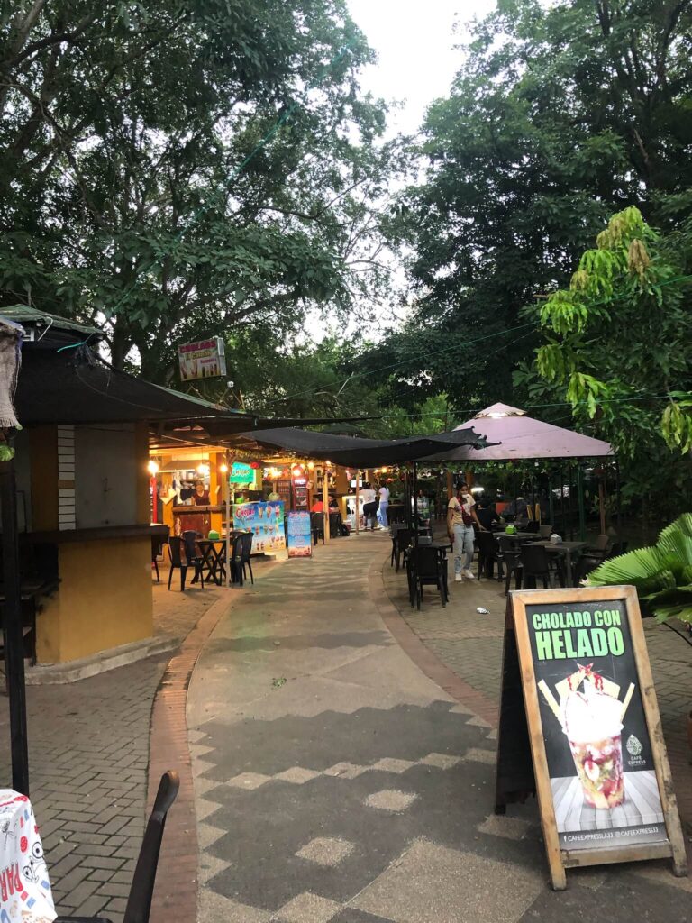 Een smal straat op asfalt dat loopt door het park heen. In het straatje zie je een paar krijtborden op de grond waarop staat wat je bij een tentje kan eten of drinken