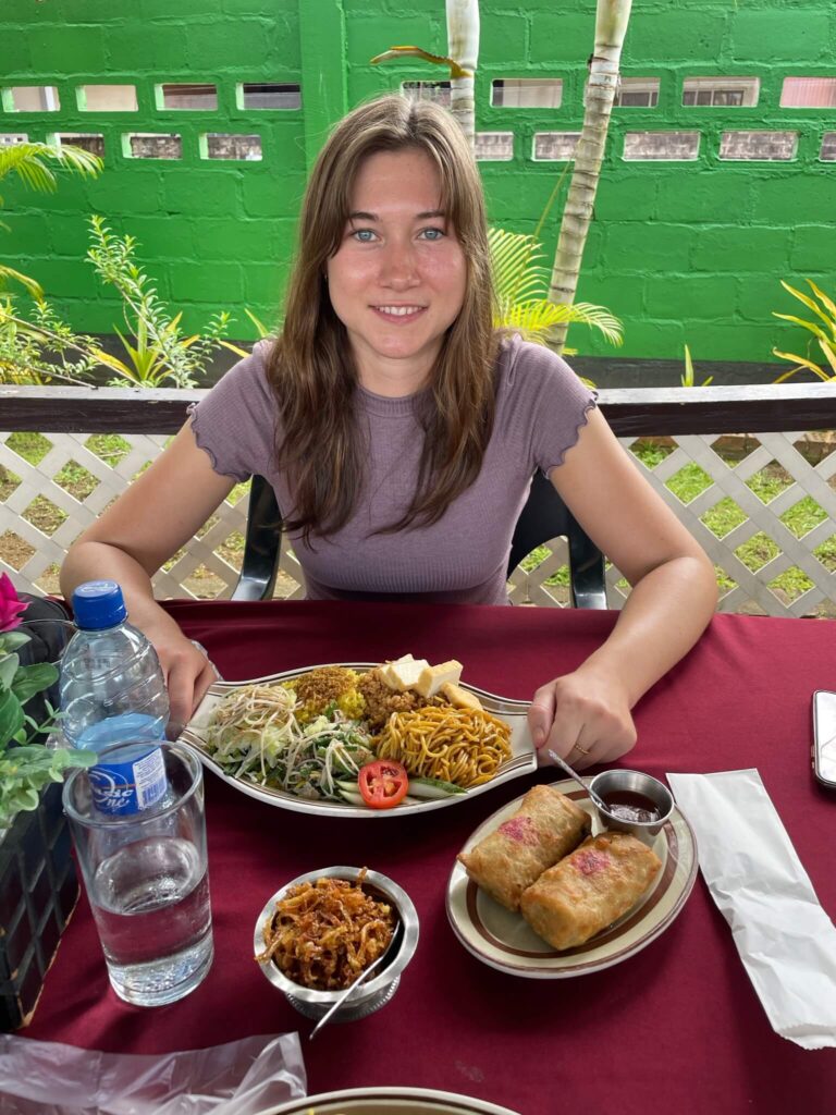 Je ziet mij aan tafel zitten en in de camera kijken. Voor mij op tafel staat een flink bord met heerlijk Javaans eten. Er staan verder ook 2 loempia's op tafel