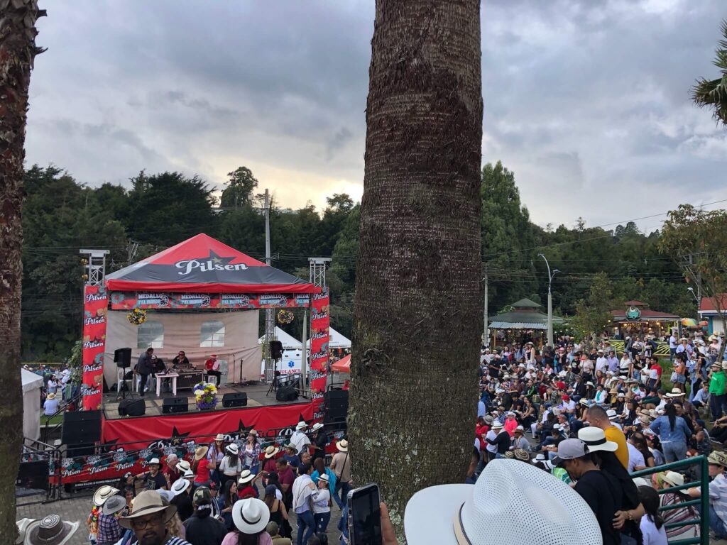 Je ziet een rood podium met daarvoor en ernaast allemaal mensen die aan het dansen zijn. Héél veel mensen dragen een witte of beige hoed