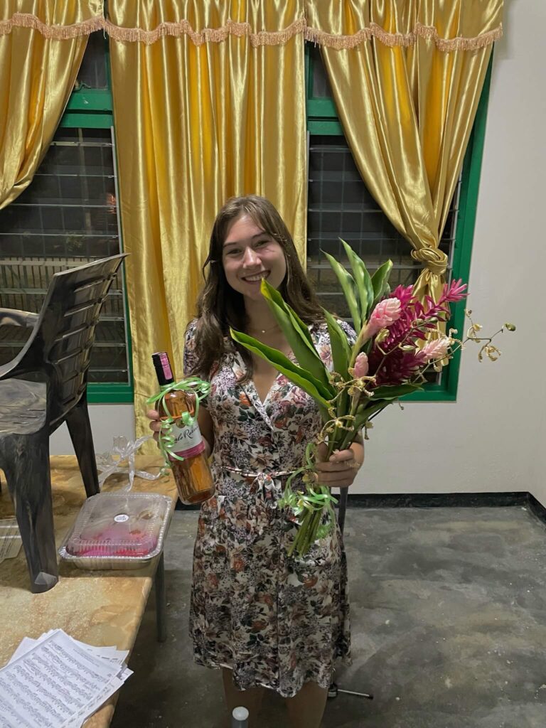 Ik poseer lachend voor de camera en heb in m'n ene hand een fles rosé vast en in m'n andere hand een bos bloemen. Links van mij staat de eettafel en daarop ligt nog een doos met donuts