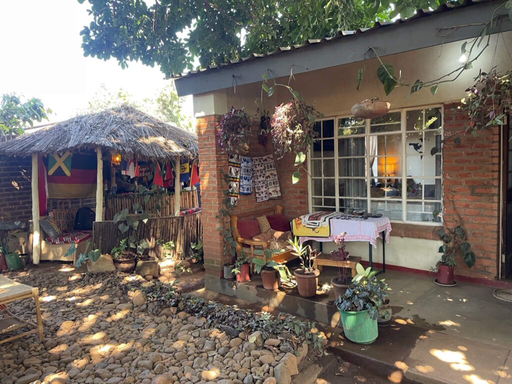 Je ziet hier de buitenkant van ons verblijf en een deel van de tuin. Voor het huis staat een tafel met wat lokale versieringen erop. Links naast het huis staat een gebouwde 'man cave': een rieten hutje met een bank eronder