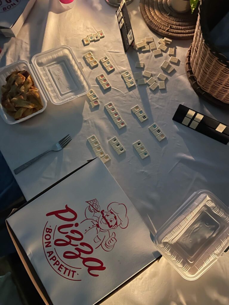 Deze foto is van de bovenkant genomen: je ziet een eettafel met daarop een wit tafelkleed, rummikub steentjes en een doos pizza en een take away bakje pasta.