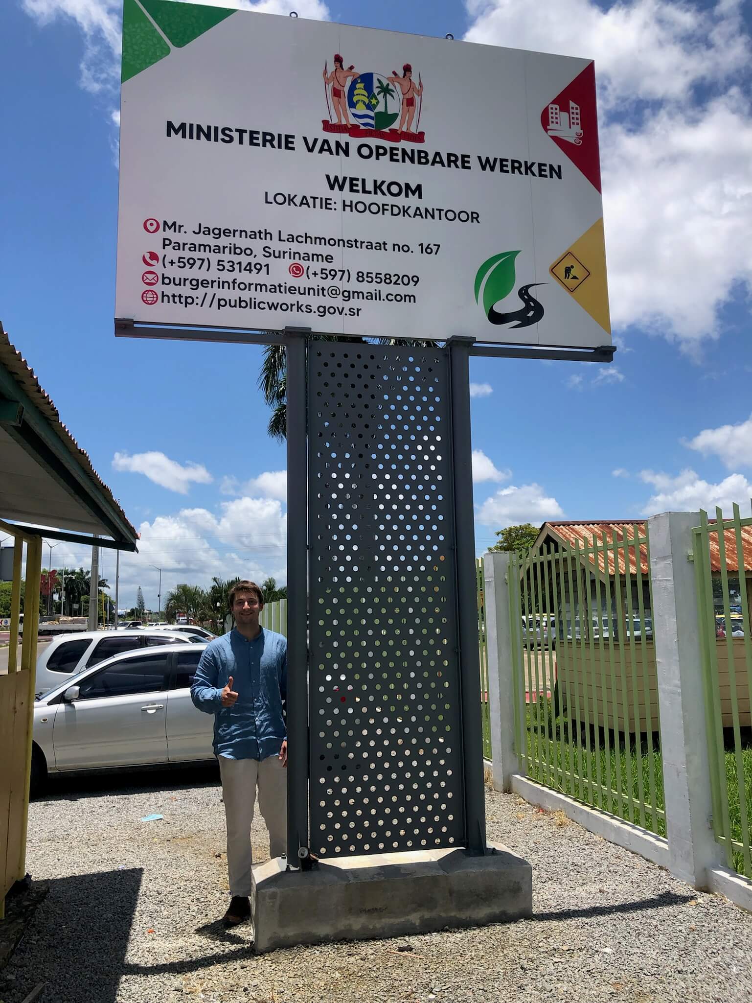 Niels staat naast/onder een groot bord waarop staat 'Ministerie van openbare werken' en 'Welkom'