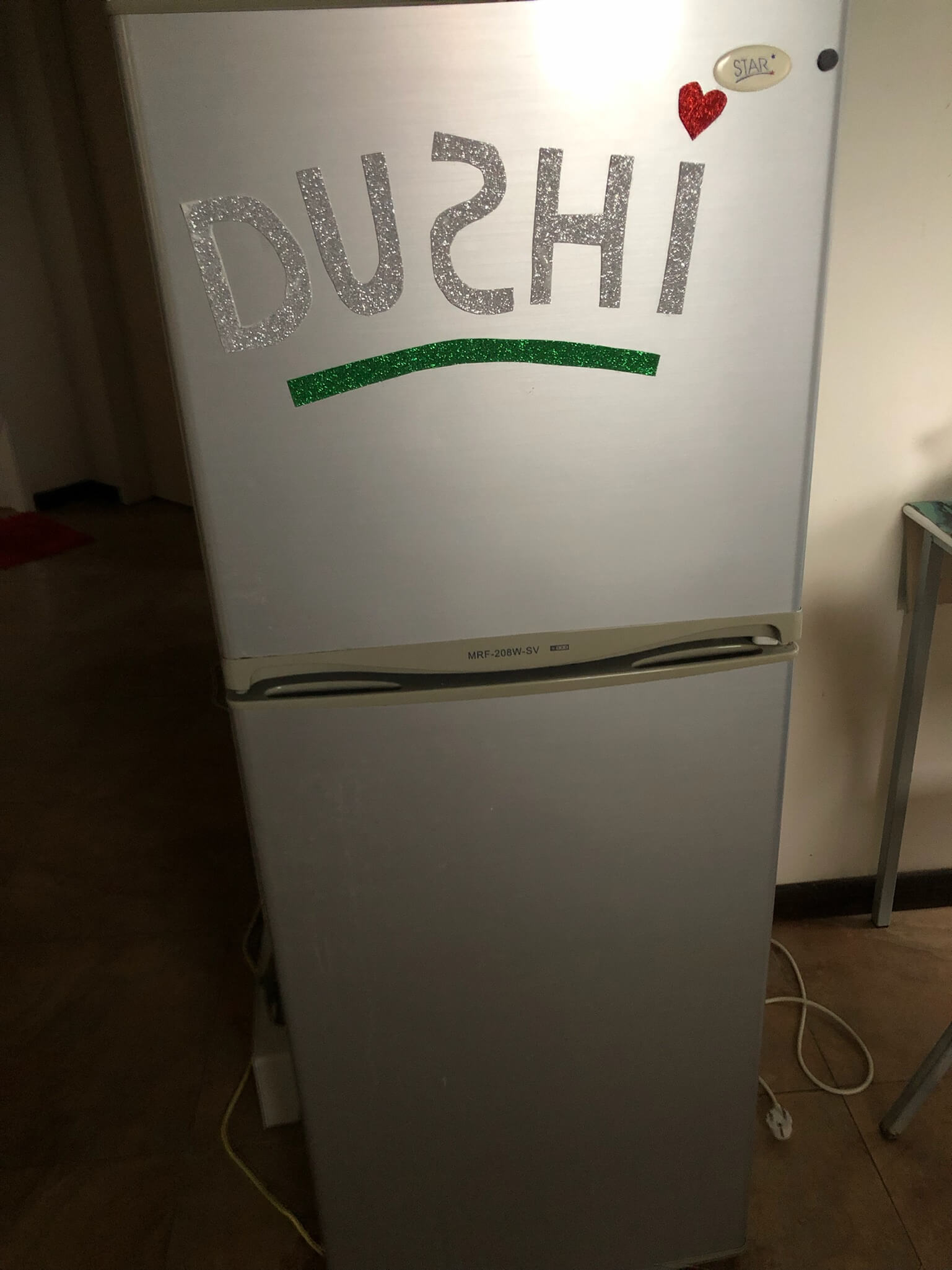 De koelkast! Een hoge grijze koelkast met daarop stickers van letters die samen DUSHI vormen