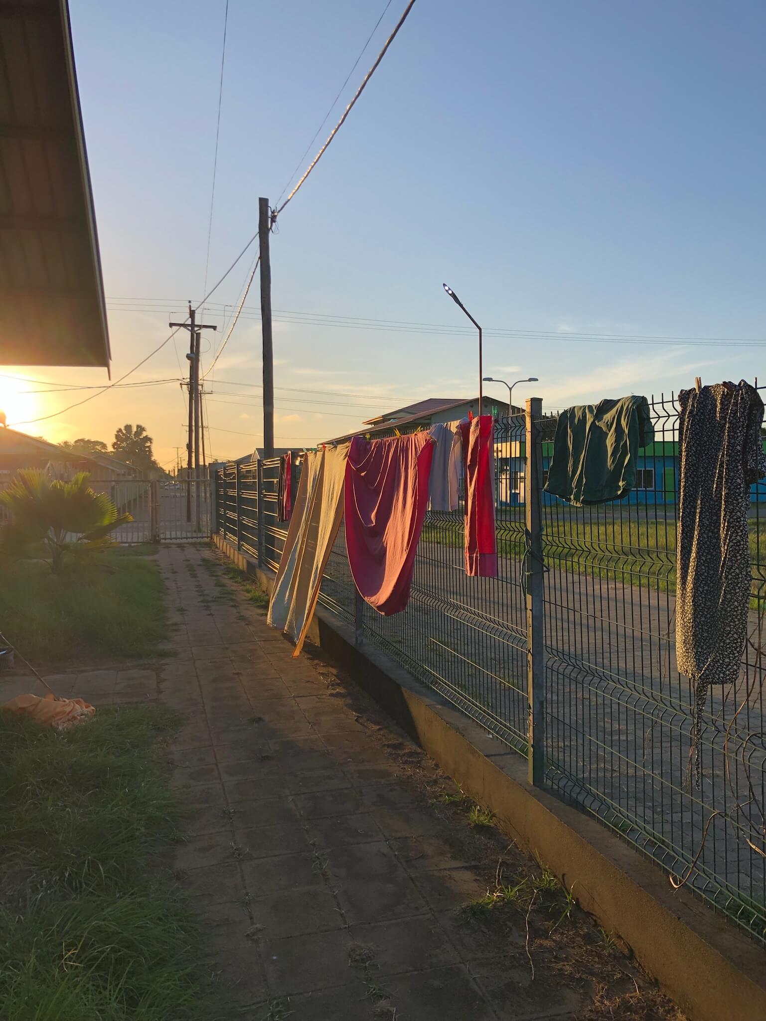 Een foto van het uitzicht van het artsenhuisje. Je ziet het hek voor het huis met daaroverheen een aantal handdoeken. In de verte zie je de zonsondergang