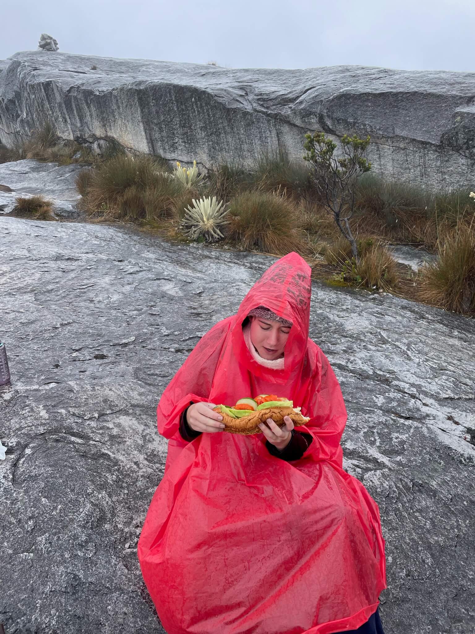 Ik zit doorweekt met een poncho aan op een grijze steen en eet een baguette. De lucht is grijs.