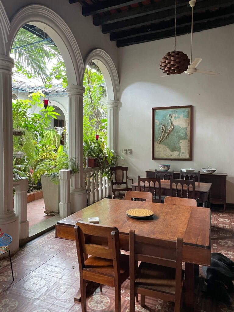 Je ziet een eetkamer met grote houten eettafel met houten stoelen eromheen, met daarachter zicht op de groene bomen en planten die in de binnentuin staan