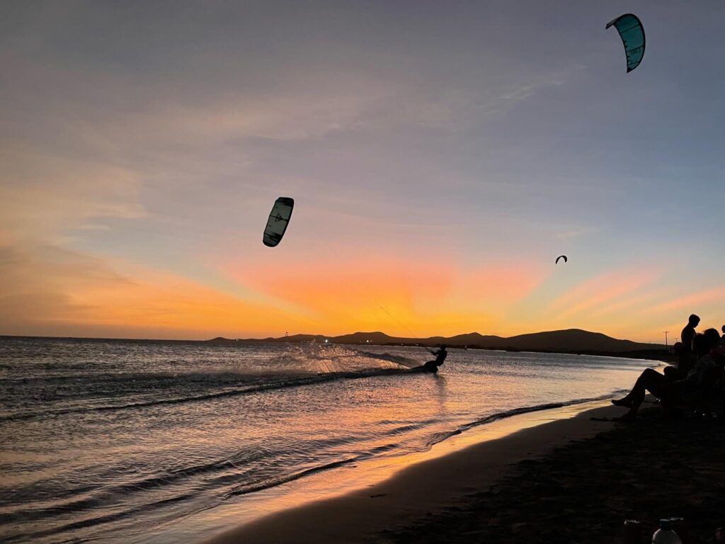 Je ziet een kitesurfer op de zee met een kite boven hem en op de achtergrond een prachtige zonsondergang