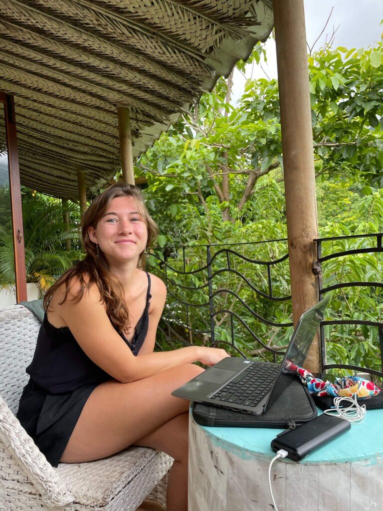 Ik zit op een stoel en heb een laptop voor me staan op een kleine tafel. Ik lach in de camera. Achter mij zijn veel groene bomen