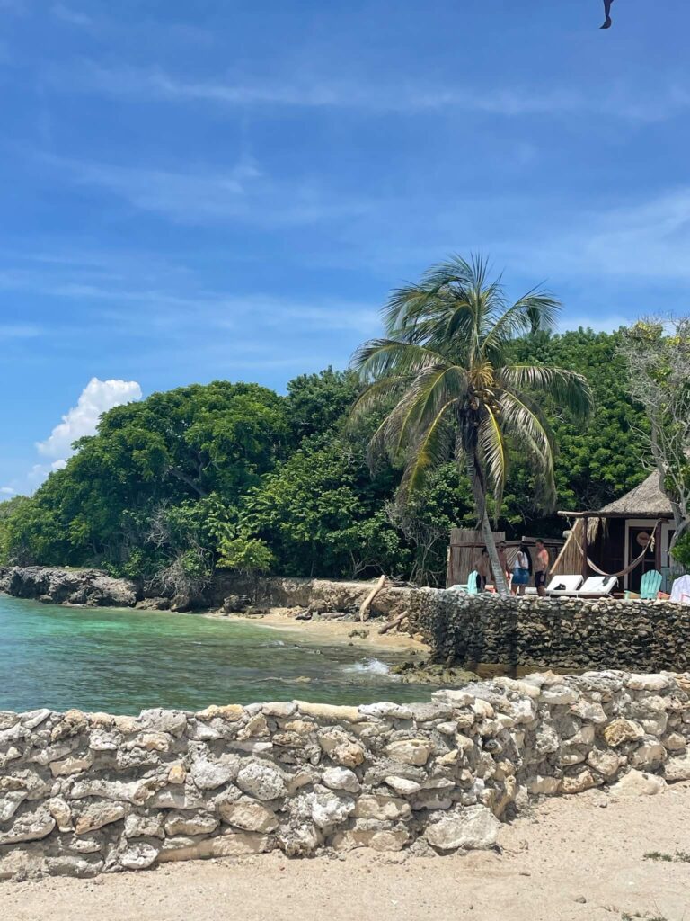 Je ziet hier de heldere zee met ernaast het strand waar ligbedjes op staan, en daarachter hoge groene bomen.