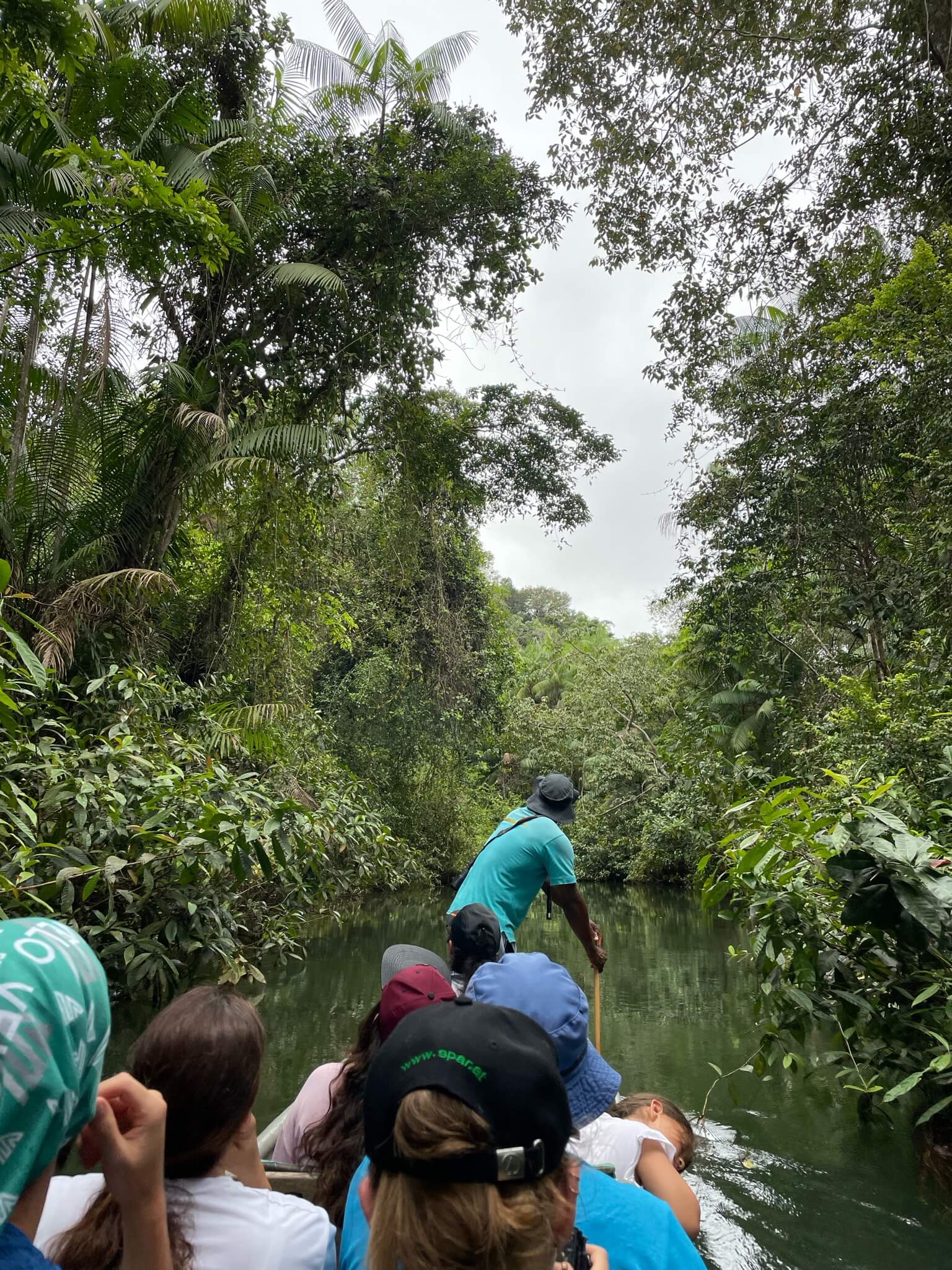 In een smalle boot zitten mensen voor ons. Voorin staat een lokale man die met een stok in het water koers probeert te varen. Om ons heen is allemaal oerwoud