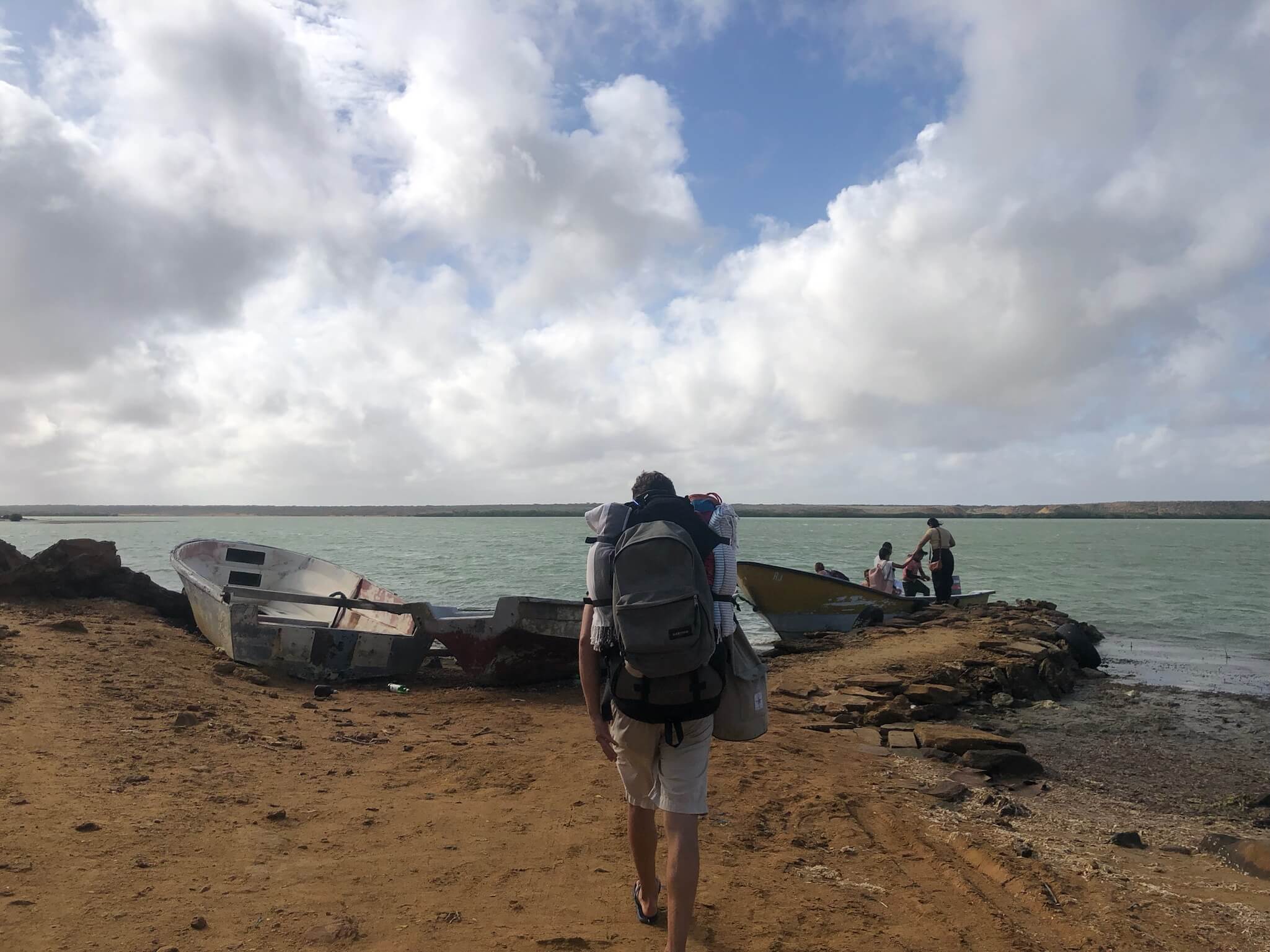 Niels loopt op het strand met een grote backpack op z'n rug. Hij loopt richting een bootje dat in de zee ligt