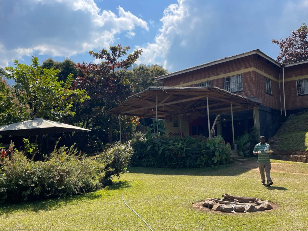 We zijn bij Blend. Je ziet hier een felgroen grasveld met op rechts het restaurant van blend in de verte. Een medewerker komt net aanlopen met een dienblad. De lucht is blauw