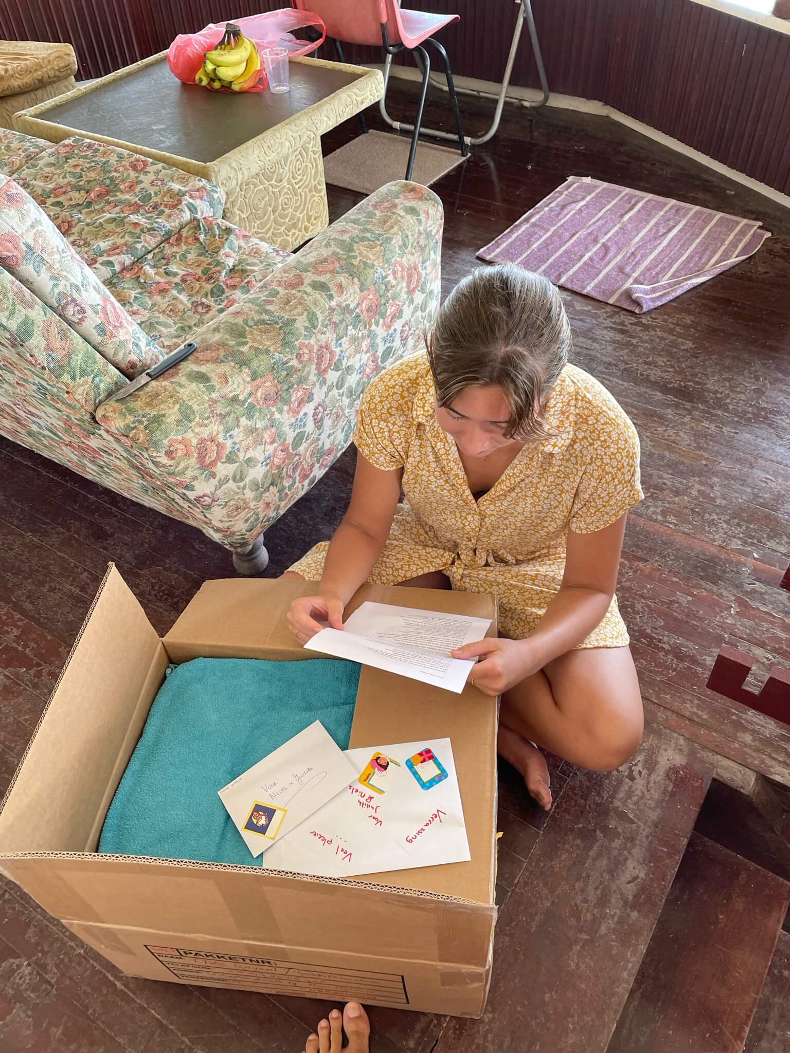 Op de grond staat de doos die we vanuit Nederland hebben ontvangen. Ik zit er in kleermakerszit naast en houd een gedicht vast