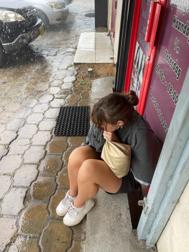 Ik zit in elkaar gedoken op een randje van een winkel onder een overkapping. Naast mij zie je een kletsnatte stoep