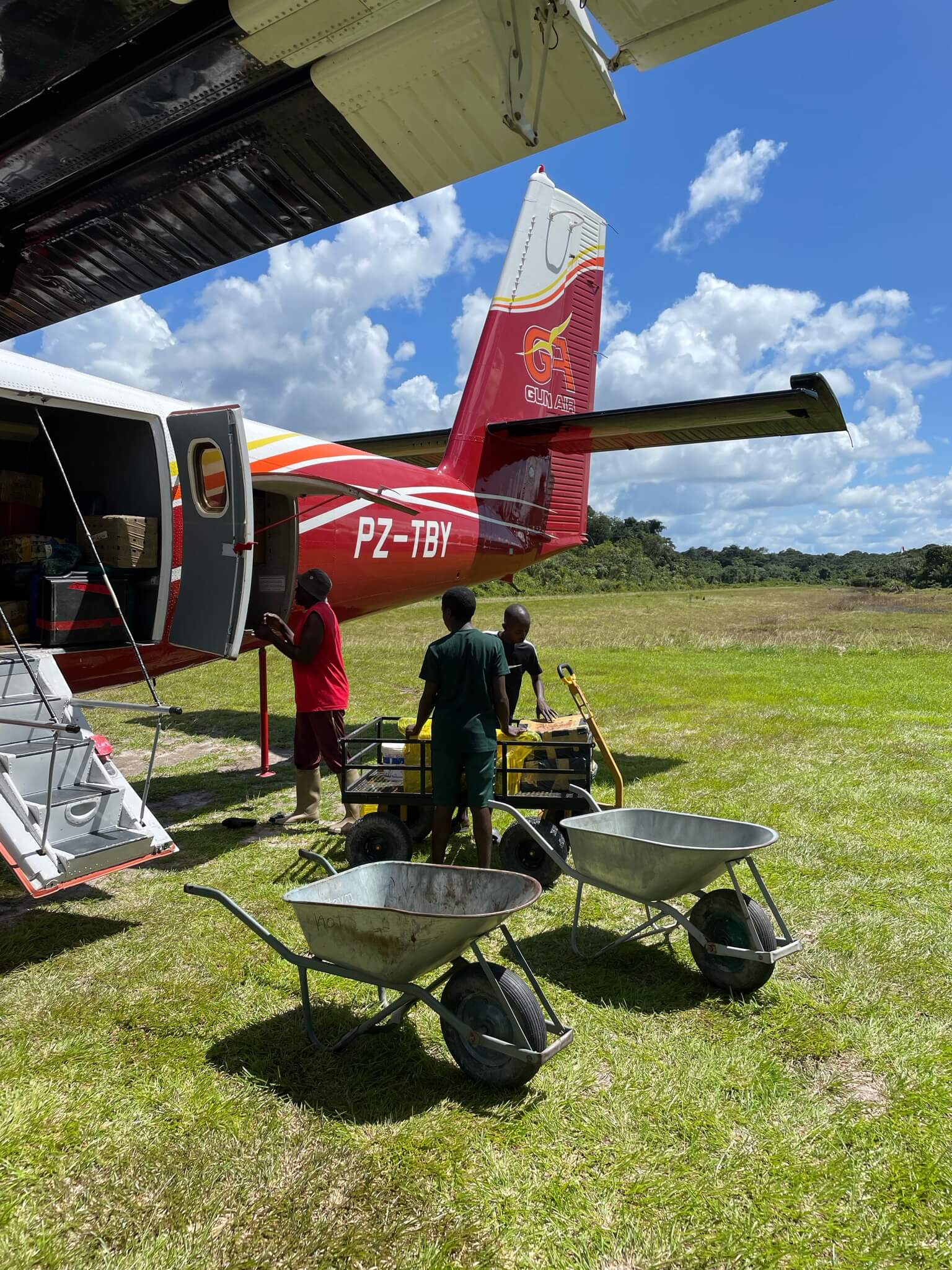 Een klein vliegtuigje staat midden op het grasveld. Naast het vliegtuigje staan 2 mannen met kruiwagens klaar om de bagage te verplaatsen