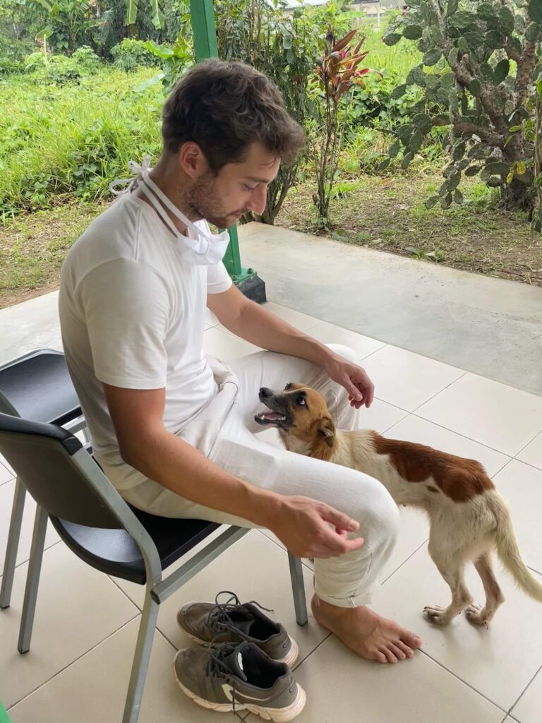 Niels zit op een stoel en tussen zijn benen staat Daisy. Ze lacht naar hem