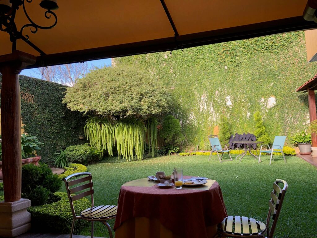 De mooie groene ontbijttuin van het verblijf: je ziet hier een ronde tafel met 2 stoelen staan met daarachter een groen grasveld en een paar enkele bomen