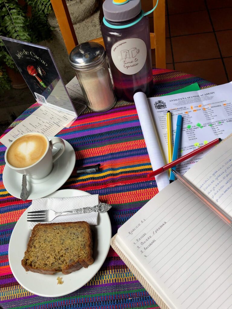 Een foto van een tafel bij een cafeetje. Op de tafel ligt mijn schrift en wat Spaanse papieren, met daarnaast een plakje bananenbrood en een cappuccino