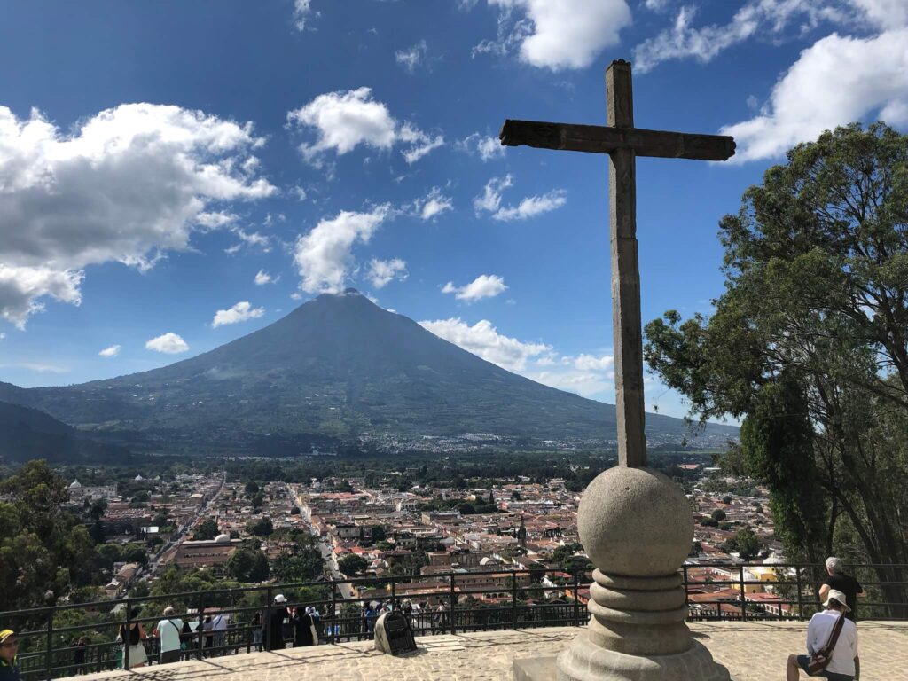 Uitzicht vanaf het uitkijkpunt 'Cerro del Cruz'. Je ziet een kruis bij het uitkijkpunt staan. Het uitzicht kijkt uit op Antigua en in de verte een vulkaan