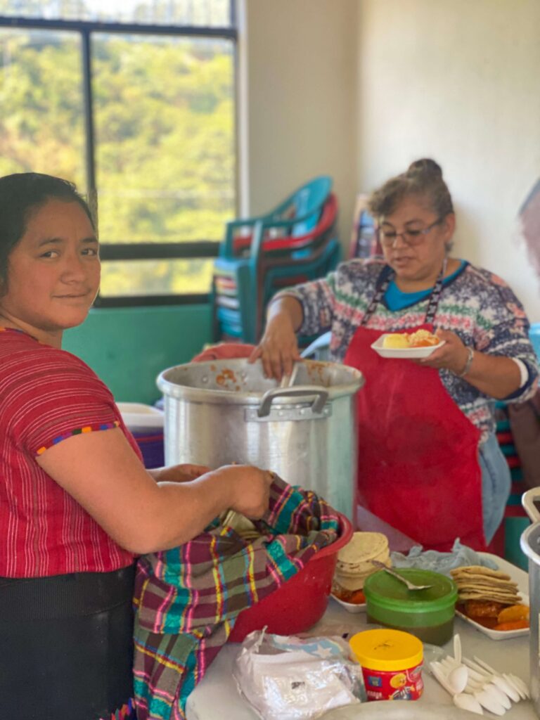 Twee lokale vrouwen staan op de foto. Eén van hen roert in een grote pan. De ander glimlacht in de camera