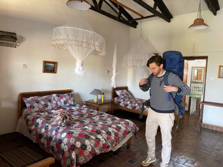 Niels staat in de slaapkamer bij Dedza met de backpack op z'n rug. In de kamer zie je twee bedden staan