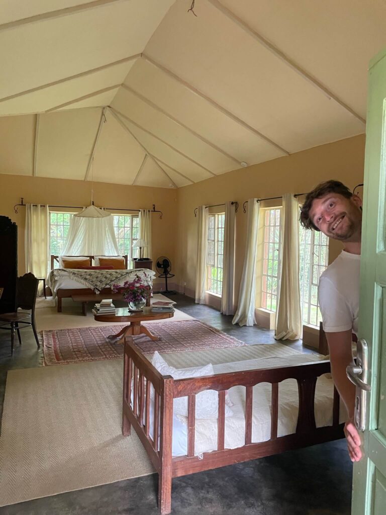 Niels doet de deur van het verblijf open en kijkt, terwijl hij achter de deur staat, lachend naar de camera. In de camera zie je het bed staan en een zitplek