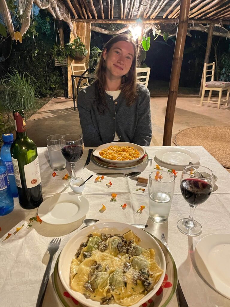 Ik poseer glimlachend voor de camera terwijl ik aan tafel zet. Op tafel staan 2 borden met pasta en 2 glazen rode wijn