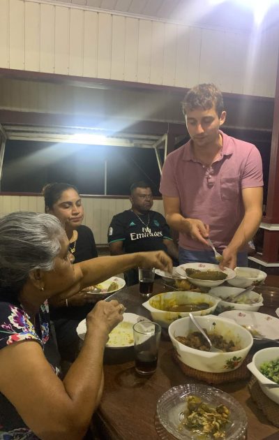 De tafel staat vol met borden met eten. Je ziet Niels en de Surinaamse familie